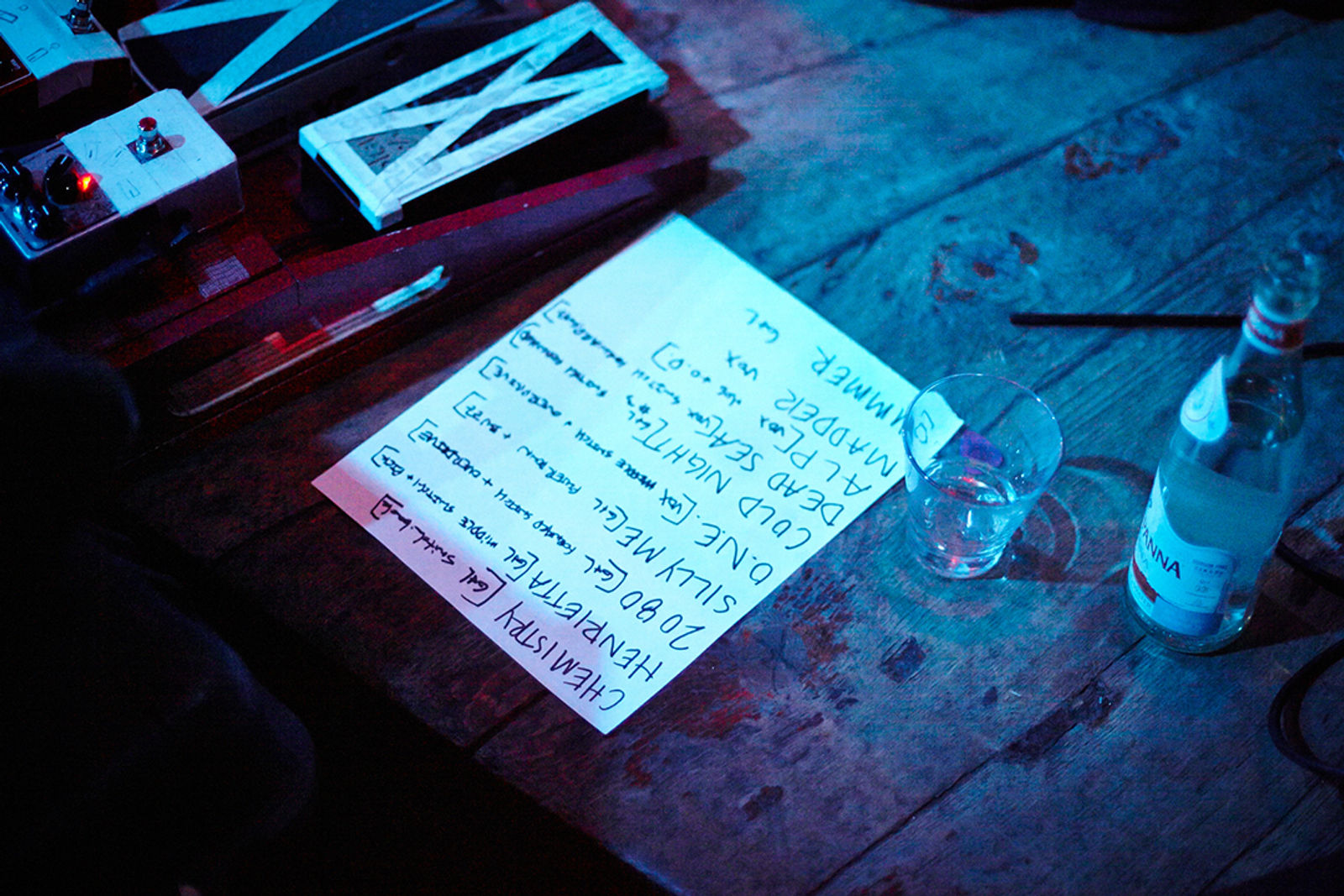 A handwritten setlist taped to a stage floor beside guitar pedals